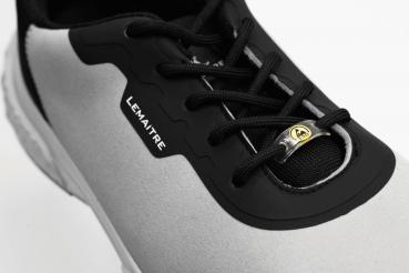 Das Bild zeigt einen sportlichen Schuh in Grau und Schwarz. Er hat eine flache Sohle, Schnürsenkel und ein auffälliges Logo. Die Materialien wirken leicht und atmungsaktiv.