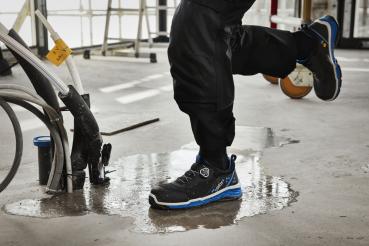 A foot stands on a wet floor, surrounded by tools and hoses. The shoe is black with blue accents, the pants are dark and protect against moisture.