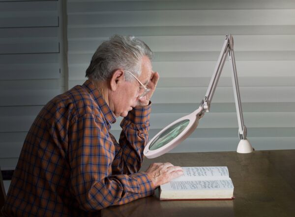 An elderly man with gray hair and a plaid shirt sits at a table, looking intently into a book. A magnifying glass and a lamp assist him in reading.