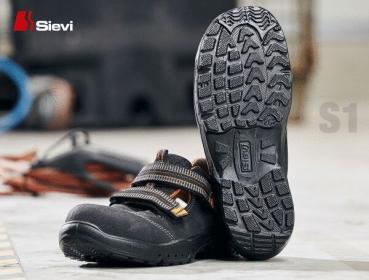 Das Bild zeigt ein Paar schwarze Sicherheitsschuhe mit einer rutschfesten Sohle. Die Schuhe haben breite Riemen über dem Fuß und sind für den Einsatz in einer industriellen Umgebung geeignet.