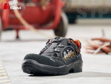 The image shows a safety shoe on a concrete floor. The shoe is black with orange accents and has a sturdy, non-slip sole. In the background, cables and a blurred machine element can be seen.