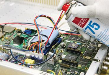 Eine Hand in einem weißen Handschuh hält eine Sprühdose über einer geöffneten Elektronikplatine. Bunte Kabel und Bauteile sind sichtbar. Der Fokus liegt auf der Reinigung oder Wartung der Technik.