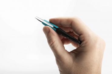 A human hand is holding a pair of tweezers. The tweezers are black with blue accents and have pointed ends. The background is white and neutral.