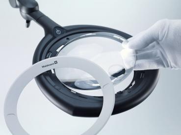 The image shows a magnifying glass with a round, clear lens. This is mounted on a black holder, while a hand wearing a white glove lifts the lens.