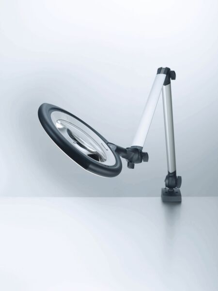 The image shows a flexible magnifying lamp with a large, round light ring. It stands on a stable base and has an adjustable arm that positions the lamp in various orientations.