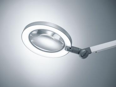 The image displays a magnifying lamp with a round, transparent glass and an integrated ring of light. The arm is flexible and allows for various positions.
