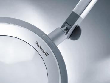 The image displays a modern magnifying glass with a large, round lens, surrounded by a slim silver frame. An adjustable arm allows for flexible positioning.