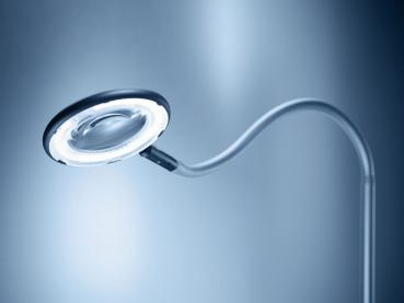 A flexible, modern reading magnifier with a round, illuminated glass lens. The arm is slim and bendable, the background appears cool and bright.