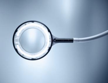The image shows a large magnifying glass with a round, illuminated frame. It has a long, flexible neck and is positioned in soft light. The background is bright and monochromatic.