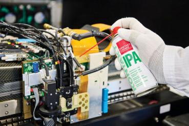 Das Bild zeigt eine Person mit einem weißen Handschuh, die eine Spraydose mit der Aufschrift "IPA" in der Nähe von elektronischen Bauteilen an einem Gerät hält. Kabel und Platinen sind sichtbar.
