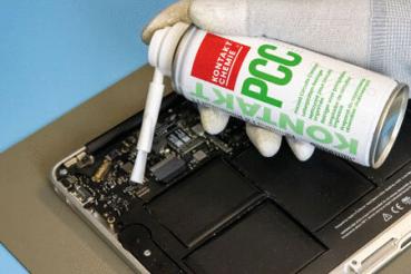 Eine Hand mit einem weißen Handschuh hält eine Spraydose über die Innenkomponenten eines Laptops. Der Sprühkopf richtet sich auf eine Platine, um Staub oder Schmutz zu entfernen.