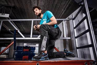 Ein junger Mann kniet auf einer Plattform in Arbeitskleidung. Er trägt ein türkises T-Shirt und schwarze Hose mit grauen Details. Im Hintergrund sind Stahlträger und eine Leiter sichtbar.