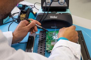 A person in a white coat is soldering with a soldering iron on a green circuit board. In the background, a Weller device is visible. The table is light blue.
