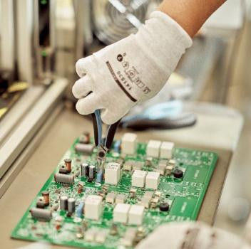 Eine Hand in einem weißen Handschuh hält eine Zange und arbeitet an einer grünen Leiterplatte mit verschiedenen Bauteilen darauf. Im Hintergrund ist eine industrielle Umgebung zu sehen.