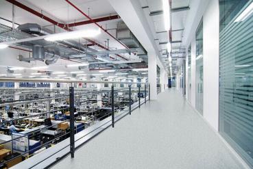 Das Bild zeigt einen modernen, hellen Büroflur mit weißen Wänden und einem gelben Boden. Auf beiden Seiten sind Büroräume und Arbeitsplätze sichtbar, mit viel technischem Equipment.