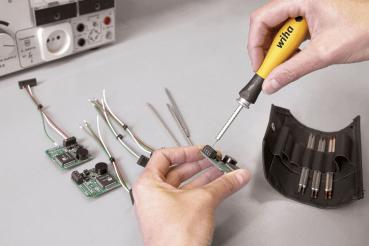 A hand holds a screwdriver over an electronic circuit board. Next to it, several cables and more circuit boards are lying around. In the background, a measuring device and a tool holder can be seen.