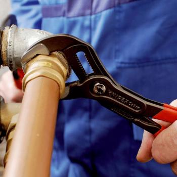 The image shows a person in blue workwear using an adjustable wrench on a joint of a copper pipe. Repair work is being performed.