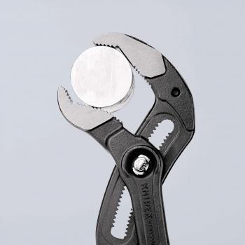 The image shows a pair of pliers with a gray, grippy plastic handle. It is holding a round, silver plate. The pliers have a straight cutting edge and a serrated area for better grip.