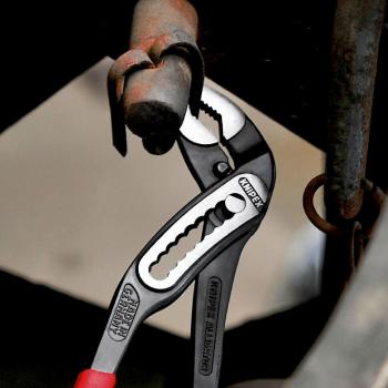 An adjustable wrench grips a thick, metallic bolt attached to an old pipe. The wrench is red and silver in color with a special clamping design.