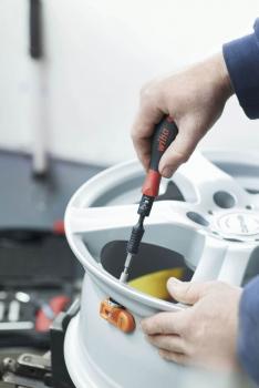 A hand holds a screwdriver tightening a screw on a silver rim. In the background, tools and a bright environment are faintly visible.