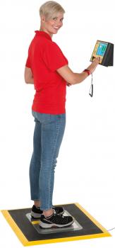 A person with short, blonde hair is wearing a red shirt and jeans. They are standing on a platform and holding a digital device in their hand. The background is neutral.