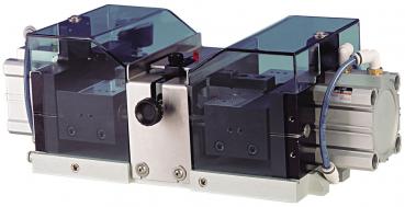 The image shows a mechanical device that has two transparent chambers, connected by a metal part. There are two lids on top and a connection for air at the bottom.