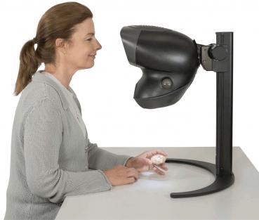 Eine Frau sitzt an einem Tisch und schaut auf ein Gerät, das wie ein Vergrößerungsgerät aussieht. Sie hält eine kleine, handliche Gegenstand in der Hand.