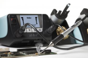 The image shows a soldering iron tool being used on a circuit board as smoke rises. In the background, there is a soldering station with digital displays.