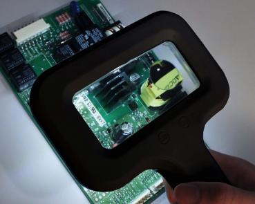 The image shows a hand holding a magnifying glass over a green circuit board. The magnifying glass illuminates an area with various electrical components, including a black element and a yellow one.
