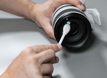 Eine Hand hält einen Objektivring einer Kamera. Mit einem Wattestäbchen wird die Linse sauber gemacht. Der Hintergrund ist schlicht und unauffällig.