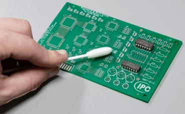 Das Bild zeigt eine grüne Leiterplatte mit verschiedenen Schaltkreisen. Eine Hand hält ein Wattestäbchen, das zur Reinigung der Platine verwendet wird.