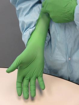 A hand is wearing a long, green rubber glove. The arm is in a light blue protective garment. The background is gray and the table is light.