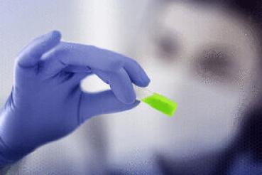 A hand with blue gloves is holding a small, brightly green object. In the background, a person with a face mask can be seen out of focus.