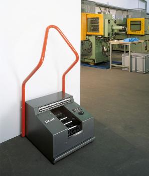Das Bild zeigt eine graue Maschinenbox mit einem großen roten Griff in Form einer geschwungenen Linie. Im Hintergrund stehen weitere Maschinen in einer Industriehalle.