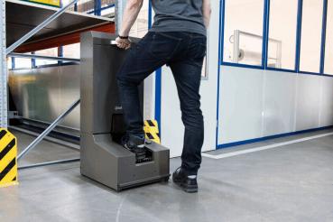 A man stands on a machine base with one foot. The machine is gray and is located in a warehouse with shelves and a white wall in the background. Safety markings are visible.