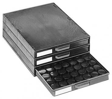 Das Bild zeigt eine dreiteilige schwarze Schubladenbox mit einem offenen Fach. In der Schublade sind viele runde, flache Objekte, die wie Deckel oder Platten aussehen.