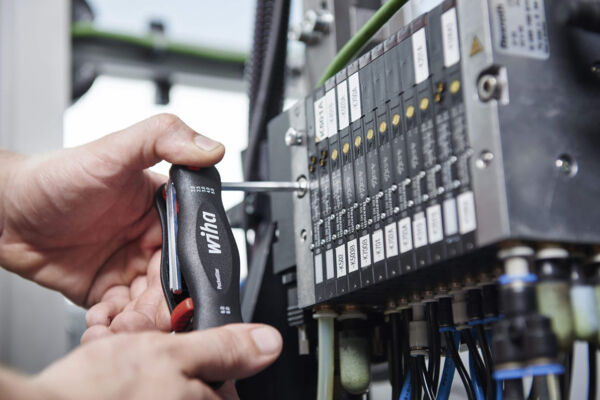 A craftsman is working with a screwdriver on a machine control. The control has multiple switches and cables that are visible. It is a technical environment.