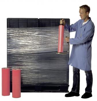 A man in a blue coat wraps a black stack of pallets with transparent film, while holding a roll in his hand. Beside him stand two pink rolls.