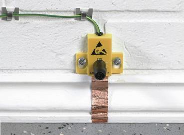 The image shows a yellow device on a wall. It features a warning symbol and is connected to a green cable. Below it, a copper strip runs along the ground.