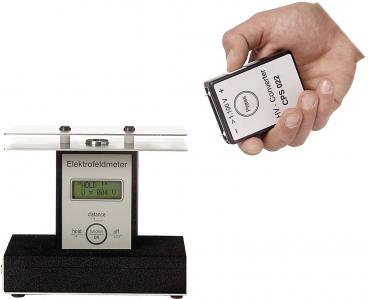The image shows an electrometer with a digital display on a black base. In one hand, someone is holding a small, rectangular device.