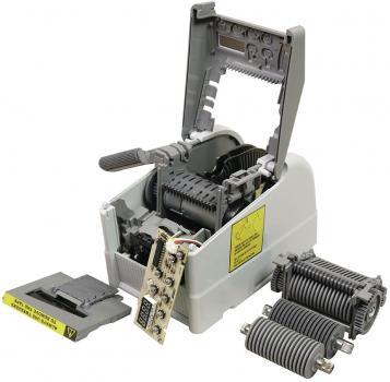 Das Bild zeigt einen offenen mechanischen Gerät mit einem grauen Gehäuse. Innen sind Zahnräder und eine Platine sichtbar. An der Seite steht ein kleiner schwarzer Block, vermutlich ein Teil des Geräts.