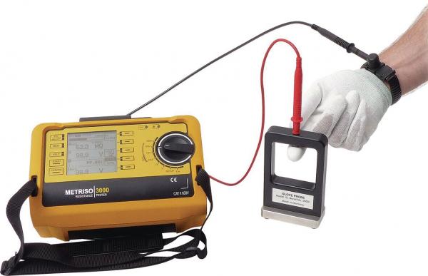Das Bild zeigt ein elektrisches Messgerät in gelber Farbe mit einem Bildschirm und Tasten. Ein Kabel verbindet es mit einem Handgerät, das eine metallische Fläche hat. Eine Hand in einem weißen Handschuh hält das Handgerät.