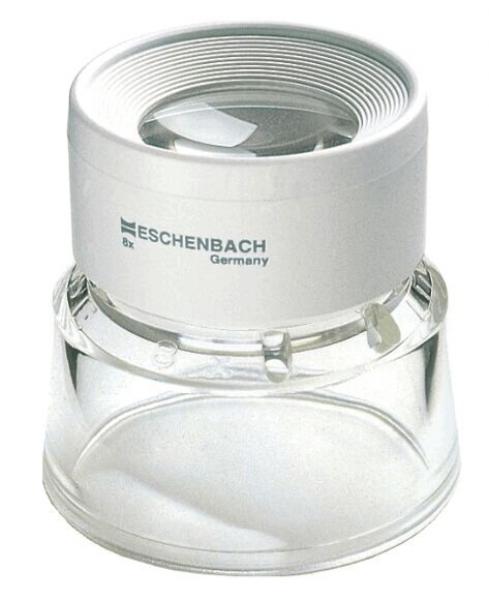 The image shows a magnifying glass in a compact, round design. At the top is a clear lens insert, below which is a slightly curved base made of transparent plastic. The surface is predominantly white.