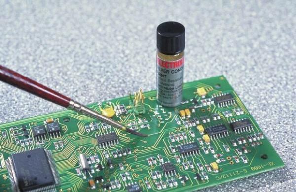 The image shows a circuit board made of green material with numerous electronic components. A red brush touches the board, while a small container stands on it.