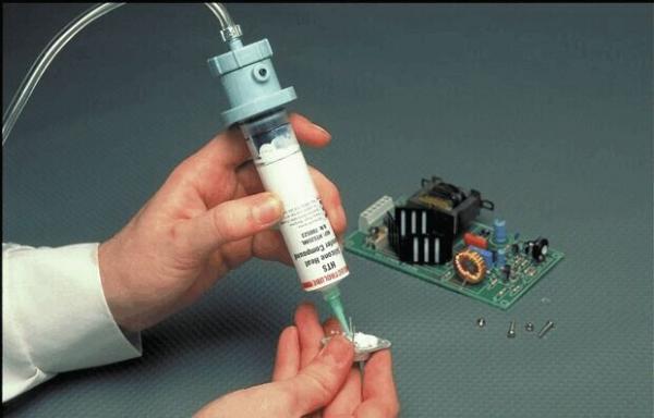 One hand is holding a syringe filled with a white material, while the other hand works on a green circuit board with various electronic components.