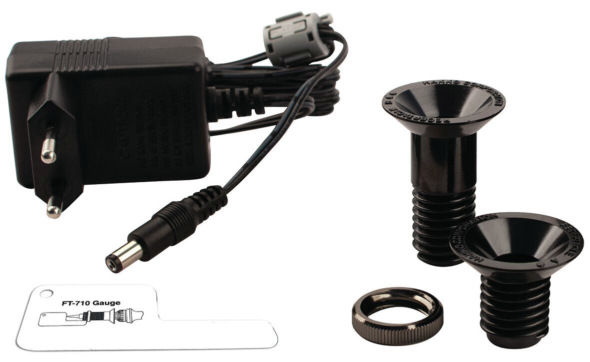 The image shows a black charger with cable and two black screw plugs. Alongside is a flat, transparent plastic measuring device.