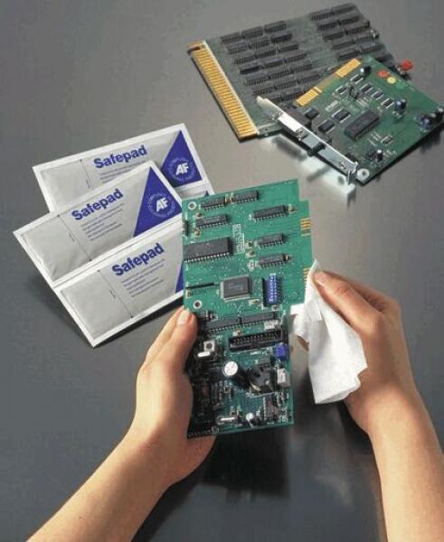 In the picture, two hands can be seen holding a circuit board and cleaning it with a cloth. In the background, there are three packs of Safepad and a second circuit board.
