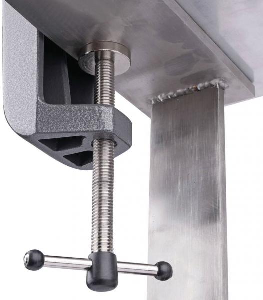 The image shows a sturdy metal clamp attached to the edge of a table. It has a long, rotating handle and a cylindrical threaded rod for fastening.