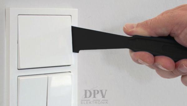 Eine Hand hält einen schwarzen Hebel, der auf einen weißen Lichtschalter zeigt. Der Schalter ist auf einer hellen Wand montiert. Der Hebel wird benutzt, um den Schalter zu öffnen.