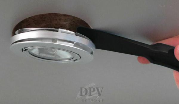 Das Bild zeigt einen Handgriff, der eine runde, silberne Deckenlampe anhebt. Die Lampe hat einen transparenten Bereich in der Mitte und ist in einen dunklen, kreisrunden Rahmen eingefasst.